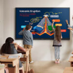 Teacher and three students point at a large digital diagram of a volcanic eruption on a classroom smartboard as classmates watch attentively.